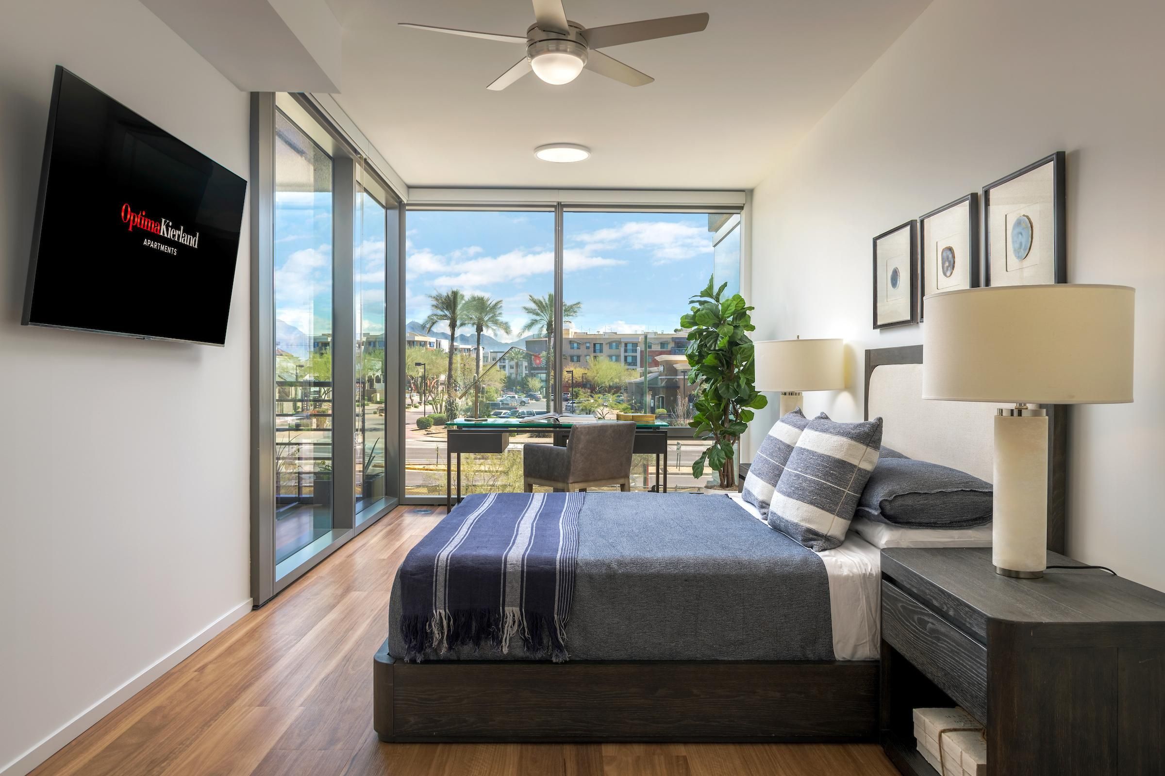 Modern bedroom with large windows, a bed with striped pillows, TV, desk, and view of a sunny outdoor area.