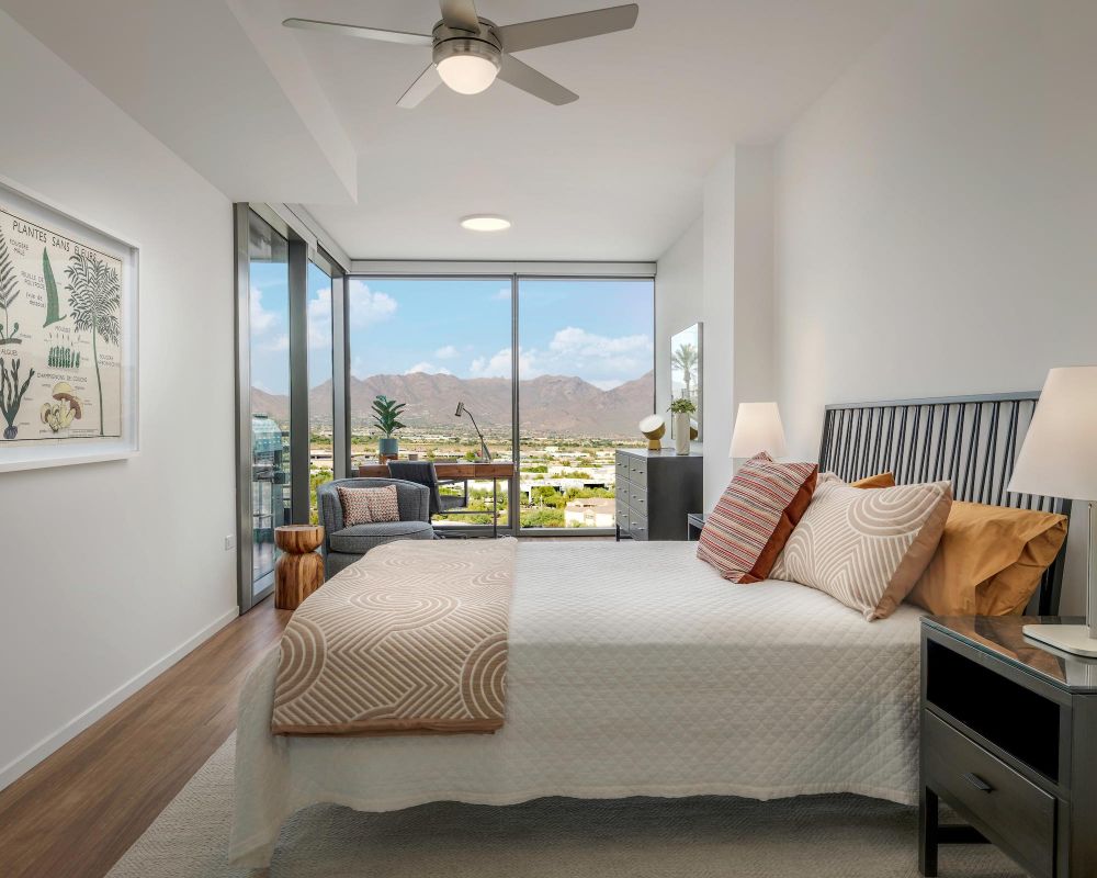Modern bedroom with large window, mountain view, bed, lamps, art on wall, and a small sitting area.