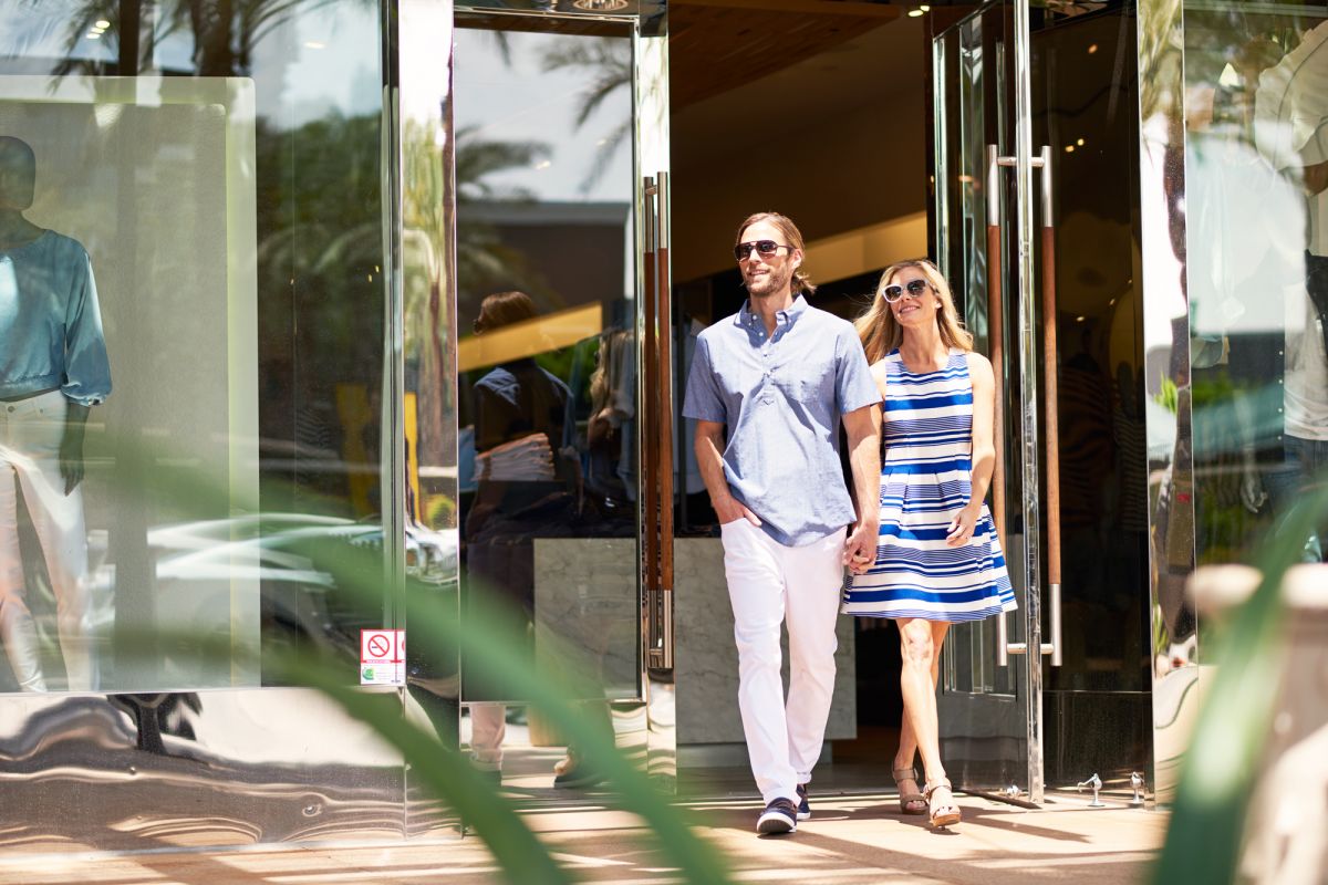 A smiling couple walks out of a store holding hands on a sunny day, enjoying the walkability and vibrant energy of urban living.