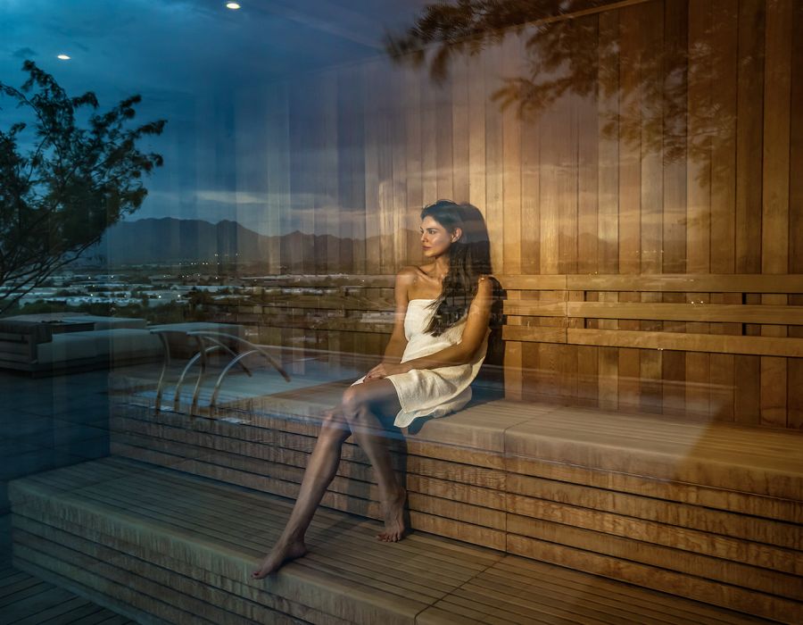 Woman in a white towel sits alone in a wooden sauna, seen through a glass window at sunset.