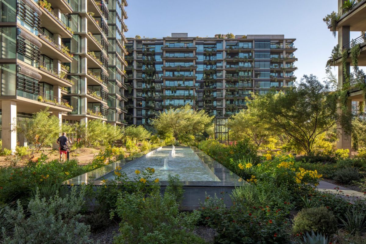 Modern apartment buildings with glass balconies overlook a lush garden and reflecting pool, elevating the Optima living community experience.