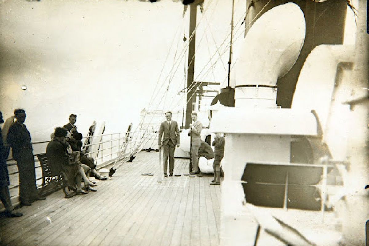 People stand and sit on the ship's deck, enjoying the ocean view and a game of shuffleboard on a cloudy day.