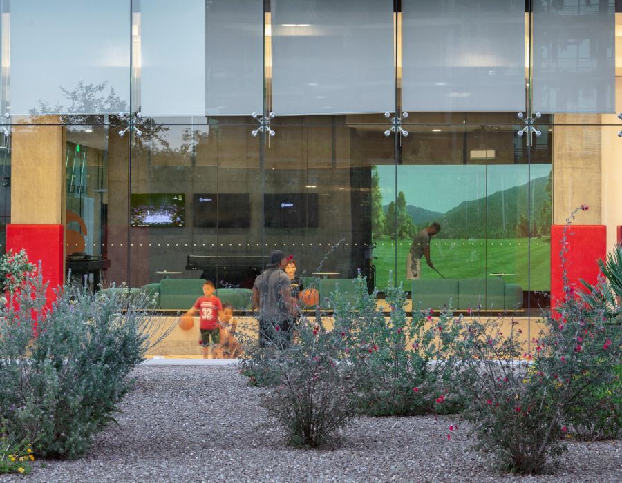 People are seen through a glass window, some watching a golf game projected on an indoor screen.