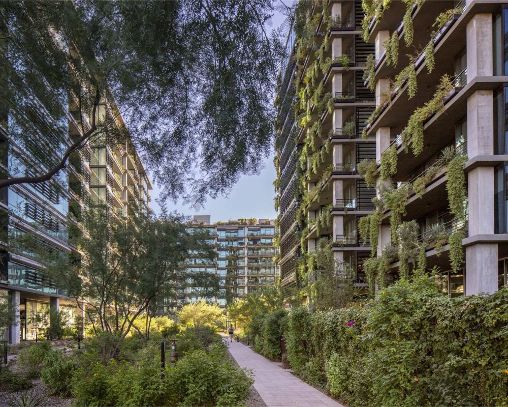 Pathway between modern buildings with balconies covered in green plants and trees, under a clear sky.
