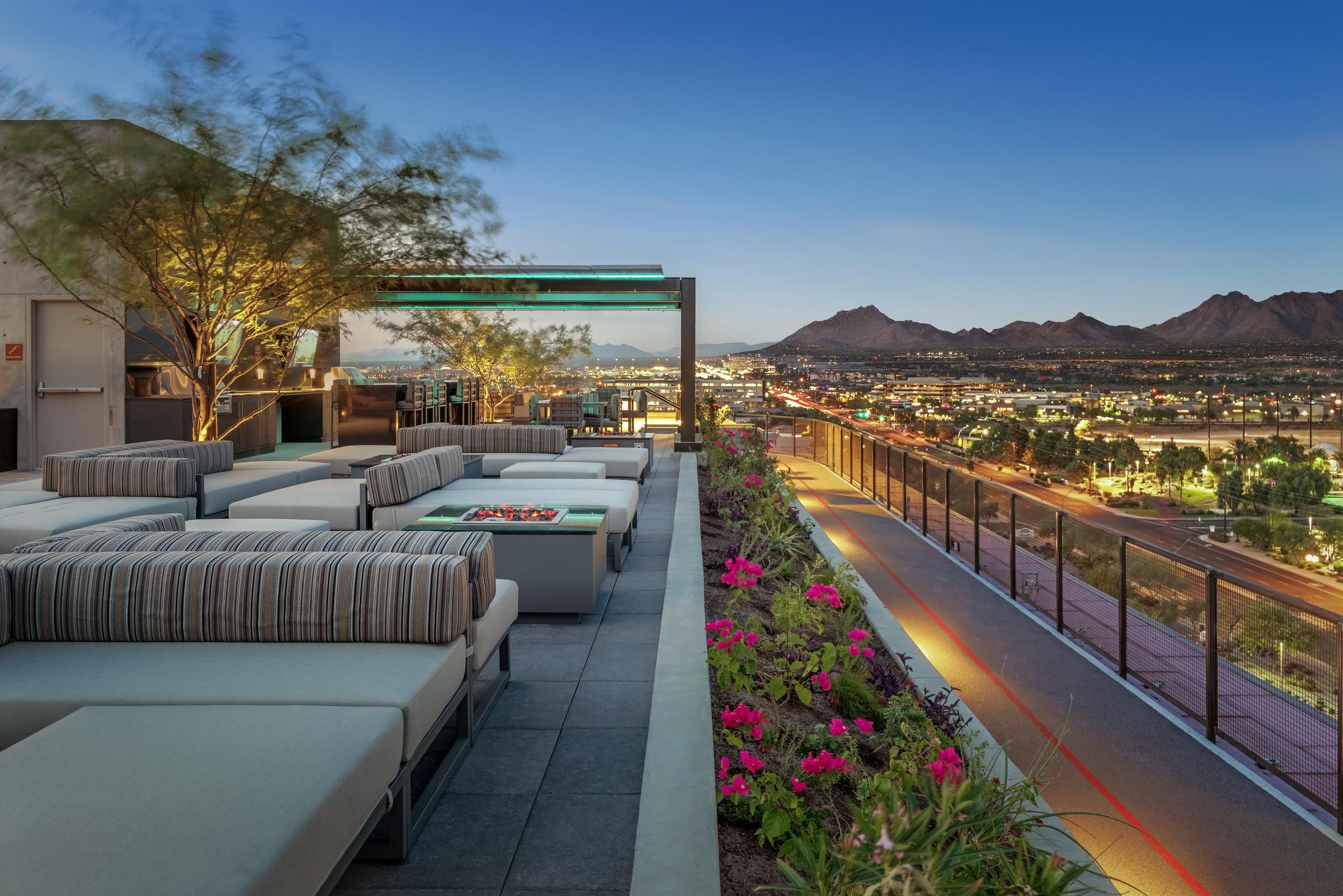 Modern rooftop lounge with striped seating, flower beds, and city and mountain views at sunset.