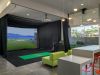 Modern room with a virtual golf simulator, green mat, clubs, lime sofa, counter, and orange stools under hanging lights.