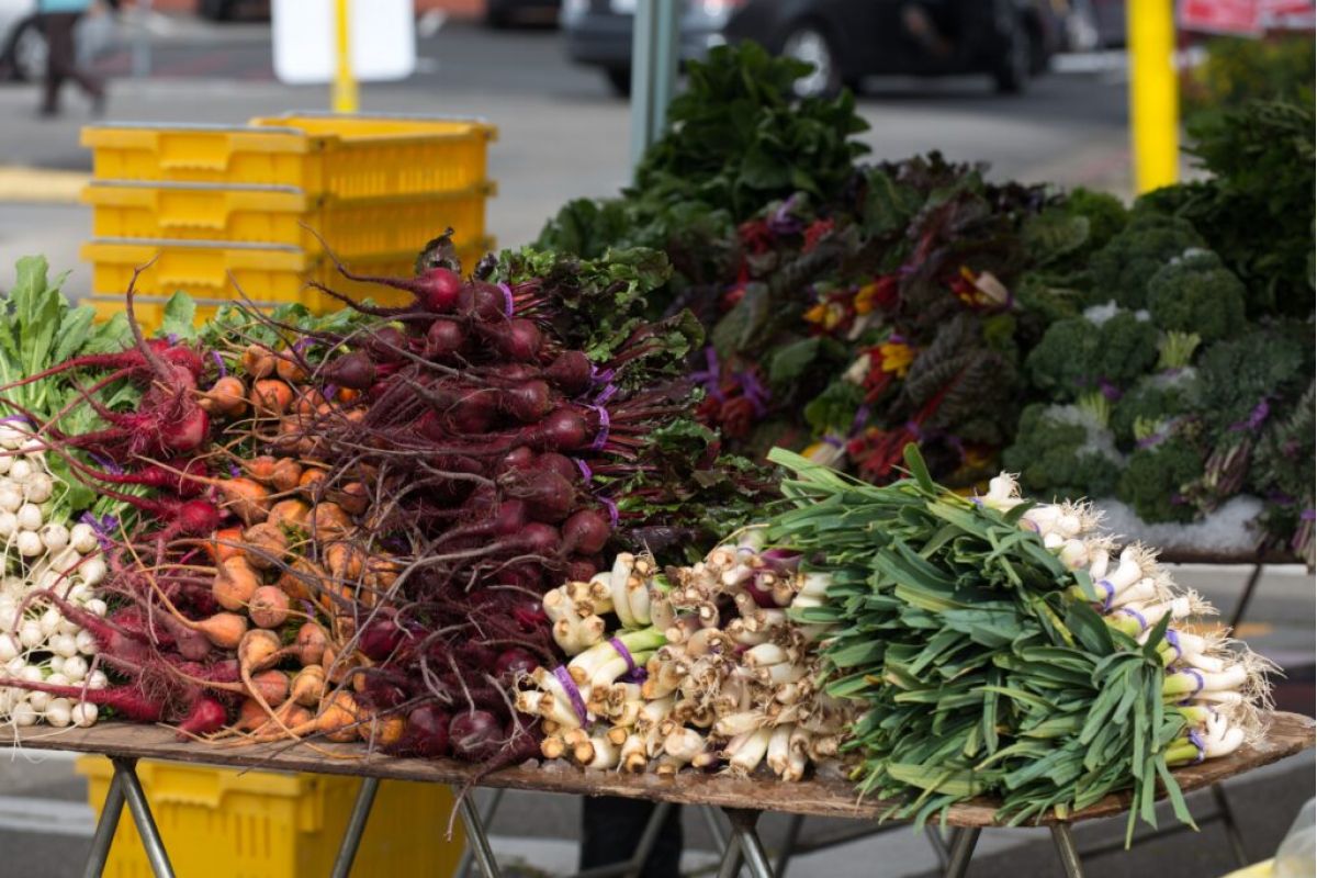 A market stand at Scottsdale Farmers Market displays fresh local root vegetables, leafy greens, and garlic in bundles.