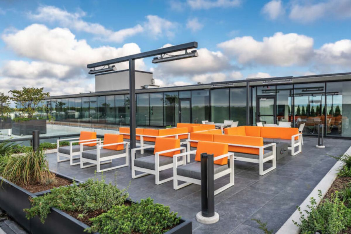 Rooftop lounge with orange seating, modern glass building, planters, and green roofs under a partly cloudy sky.