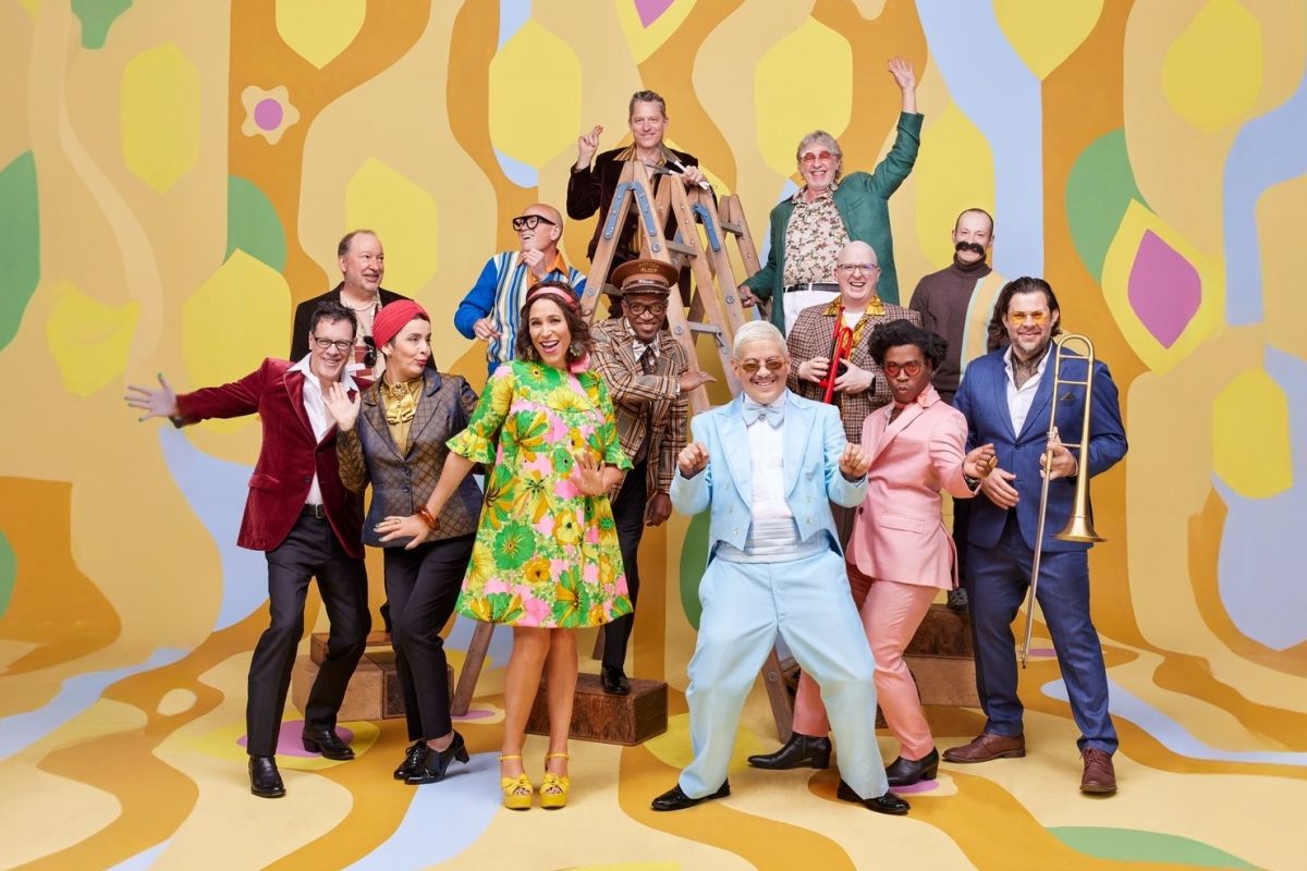A cheerful group in colorful retro outfits poses on steps, celebrating music with a vibrant, patterned backdrop for the Arizona Musicfest 2024-25 Season.