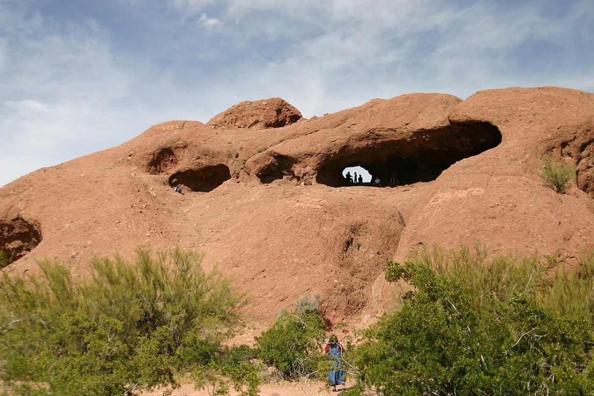 Reddish rock formation with large openings, people inside, and desert vegetation—perfect for hiking along Papago Park trails.