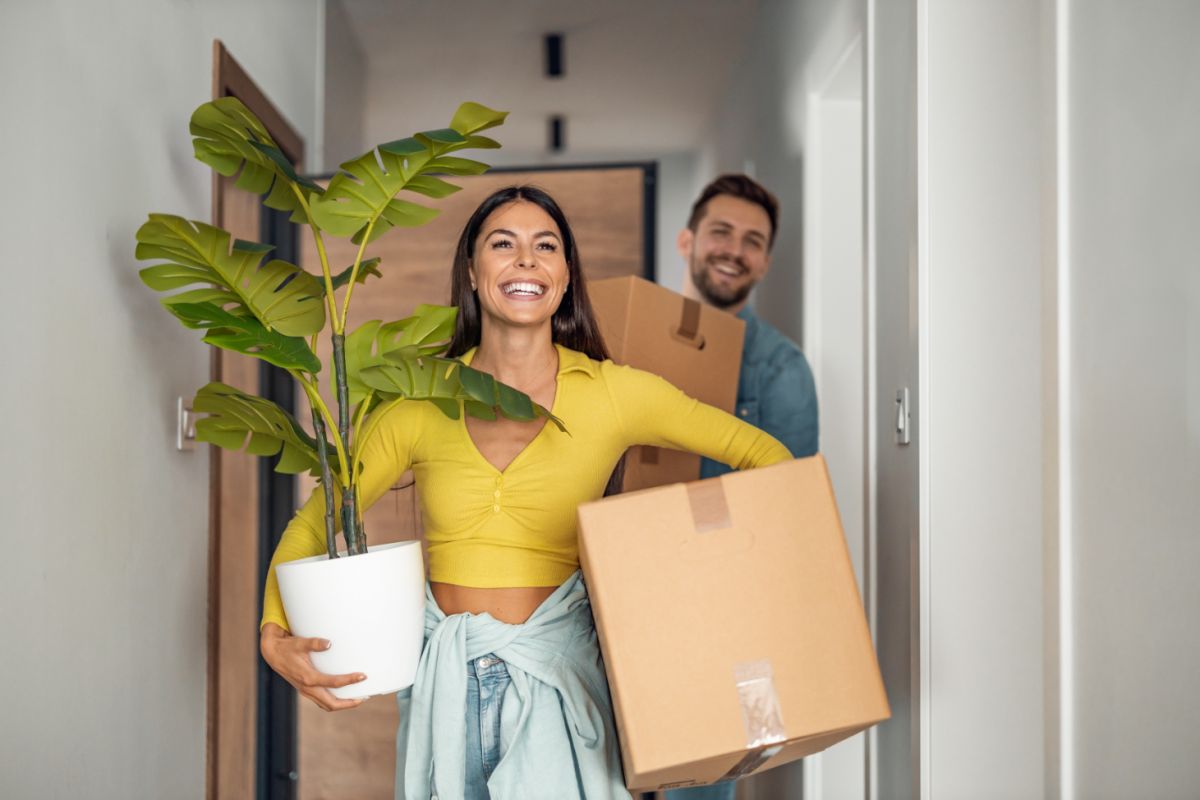 Smiling woman carrying a plant and box, followed by a man with a box, moving from house to apartment with ease.