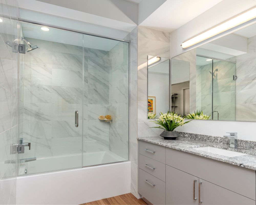 Modern bathroom with a glass shower, marble walls, granite countertop, and a vase of flowers by the sink.