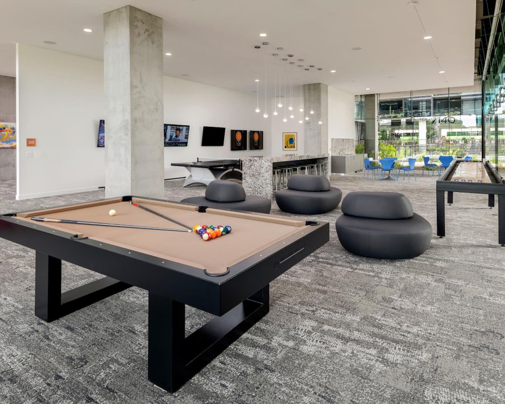 Modern game room with a pool table, lounge chairs, wall art, and large windows overlooking a courtyard.