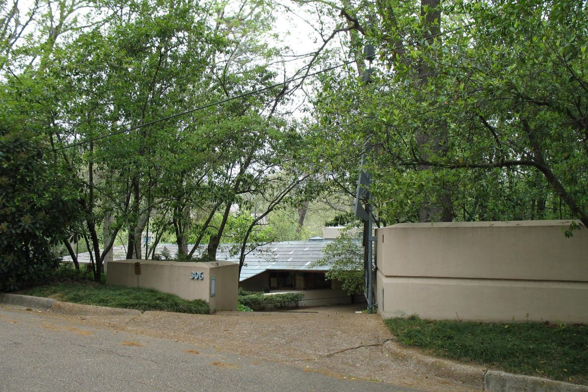 Modern house with beige walls and large windows, embracing the legacy of modernism, partially hidden by green trees and shrubs.
