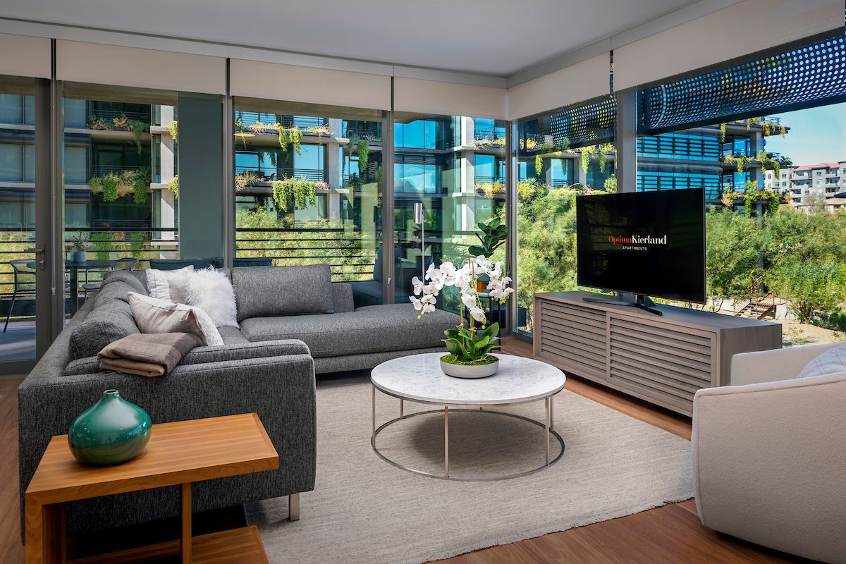 Modern living room with a gray sofa, marble table, and TV at Optima Kierland, featuring biophilic design and views of lush greenery.