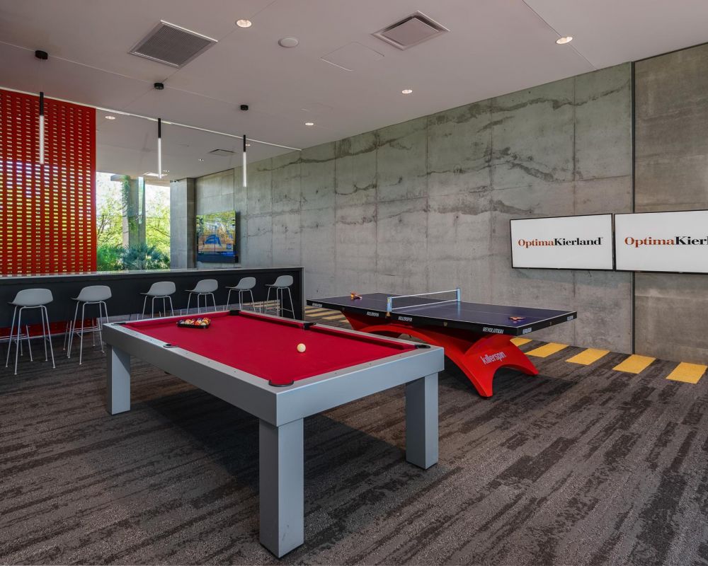 Modern game room with a red pool table, ping pong table, bar stools, and two wall-mounted TVs.