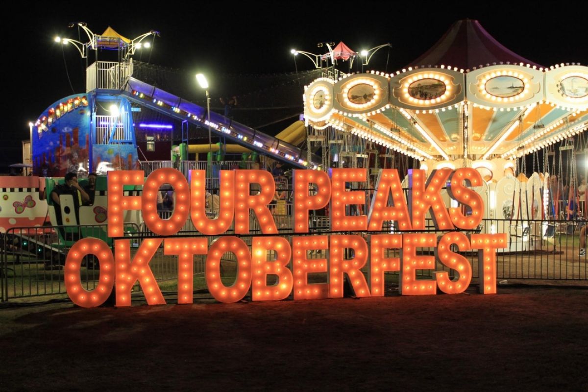 Brightly lit "Four Peaks Oktoberfest" sign at Tempe Beach Park with carnival rides glowing at night in the background.
