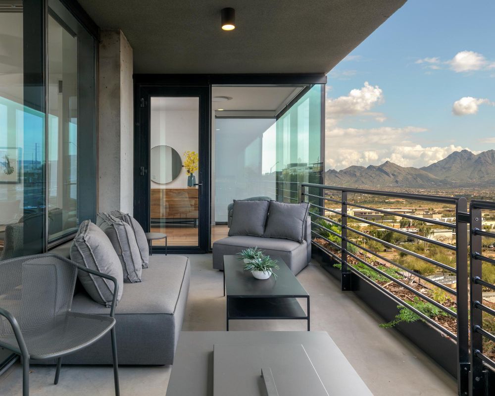 A modern balcony with gray furniture overlooks mountains and a cityscape under a partly cloudy sky.