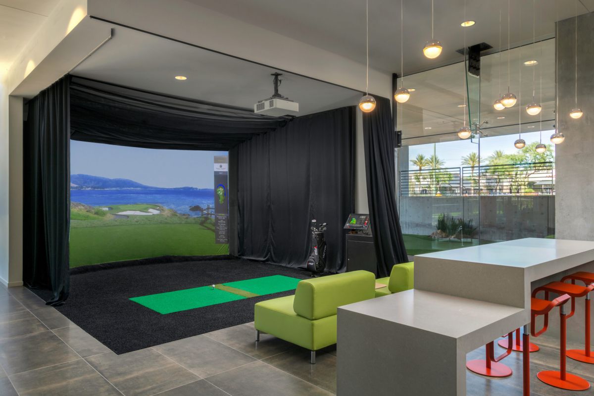 Modern room with a virtual golf simulator, green mat, clubs, lime sofa, counter, and orange stools under hanging lights.