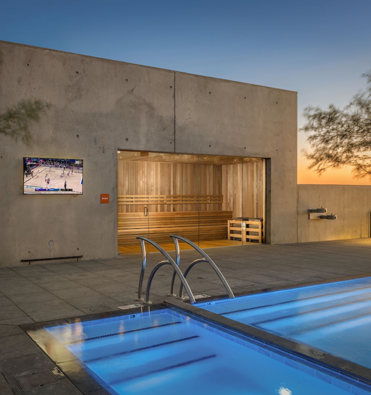 Modern rooftop at Optima Kierland Apartments with a glowing pool, sauna for health benefits, TV showing sports, and trees at sunset.