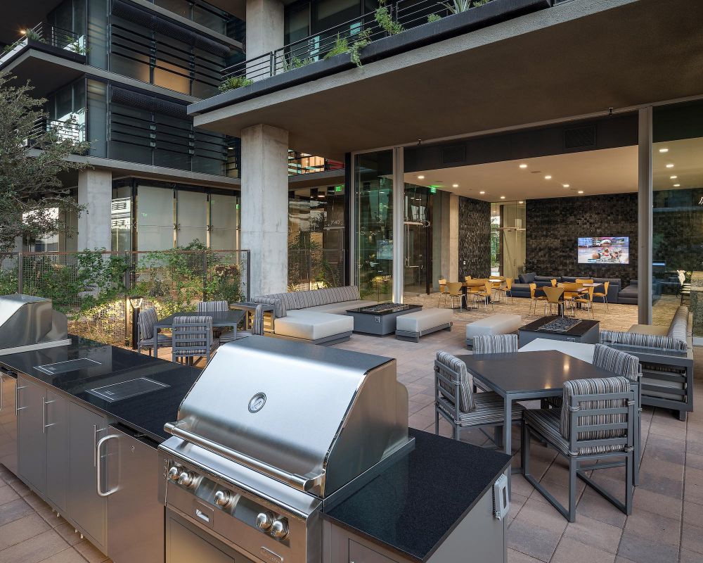 Outdoor lounge with grills, dining tables, seating areas, and a view into a modern indoor common room with TV.