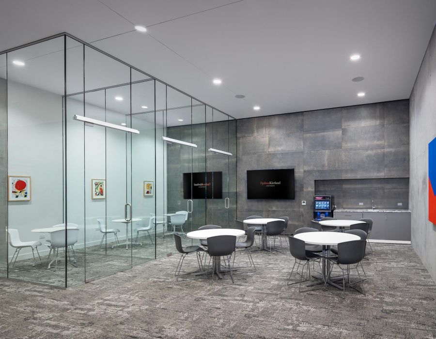 Modern office break room with round tables, gray chairs, glass-walled meeting room, and wall art.