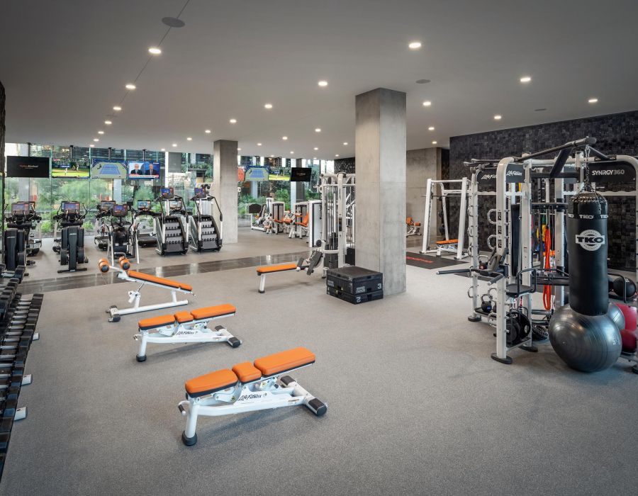 Modern gym with orange and white exercise machines, large windows, and TVs mounted on the wall.