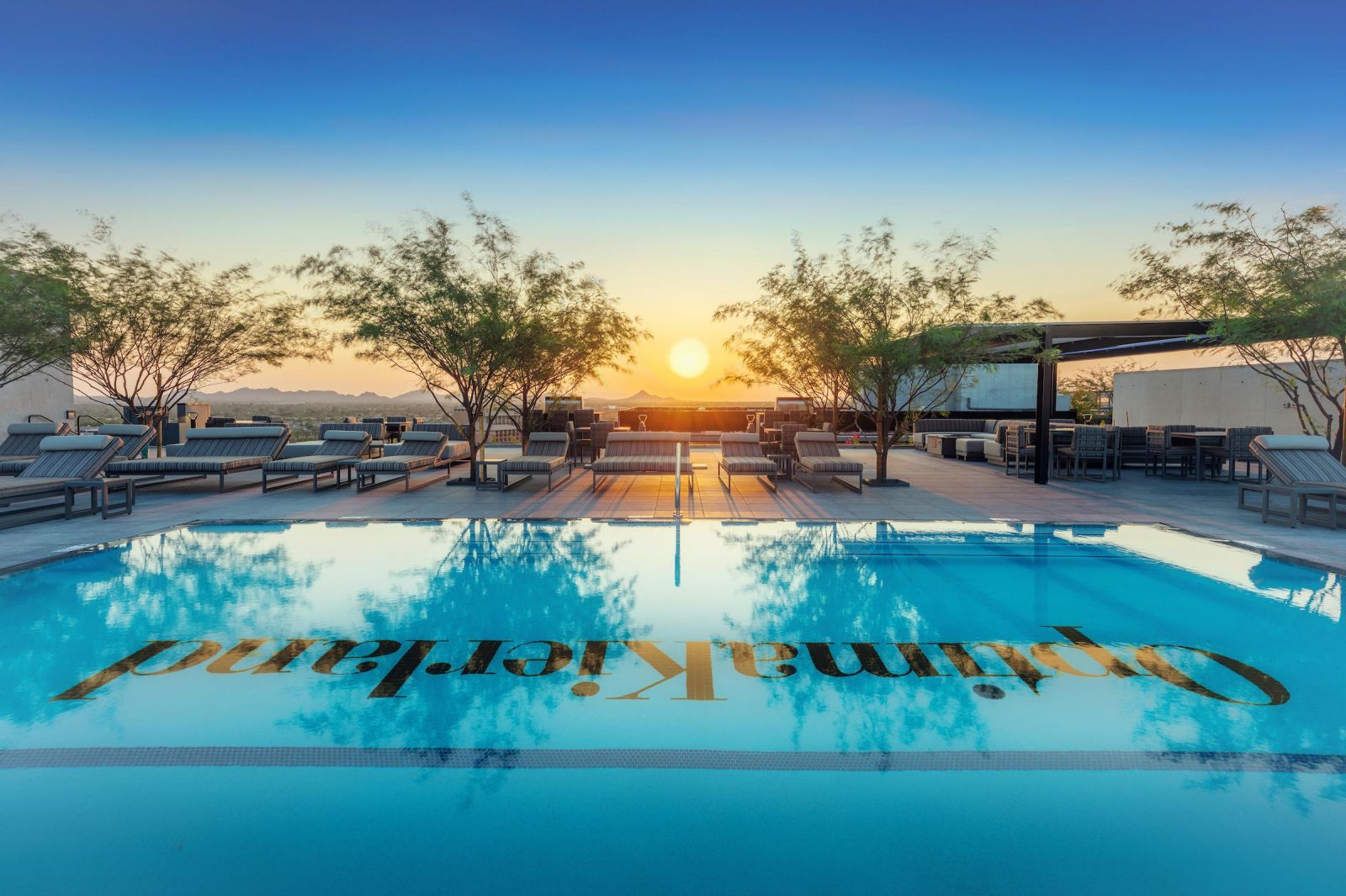 Rooftop pool with lounge chairs, overlooking a sunset-lit mountain and cityscape at Optima Sonoran Village apartments.