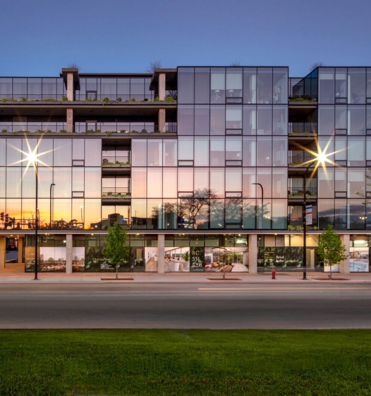 Modern glass architecture reflecting sunset, with trees and street in the foreground, bathed in natural light.