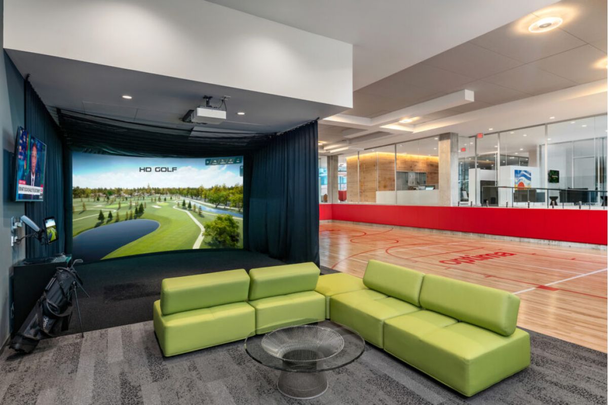 A golf simulator and green couches sit beside an indoor basketball court in this Optima Communities luxury home.