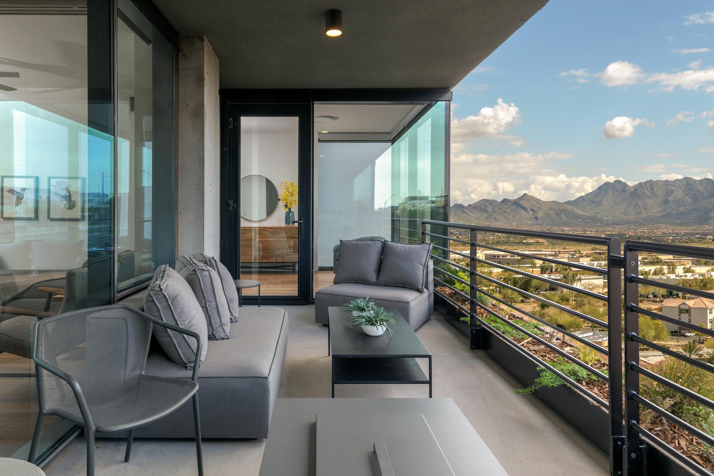 A modern balcony with gray furniture overlooks mountains and a cityscape under a partly cloudy sky.
