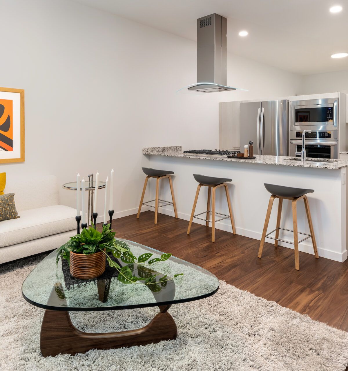Modern open-plan living room and kitchen with spotlight lighting, a sofa, bar stools, glass coffee table, and plants.