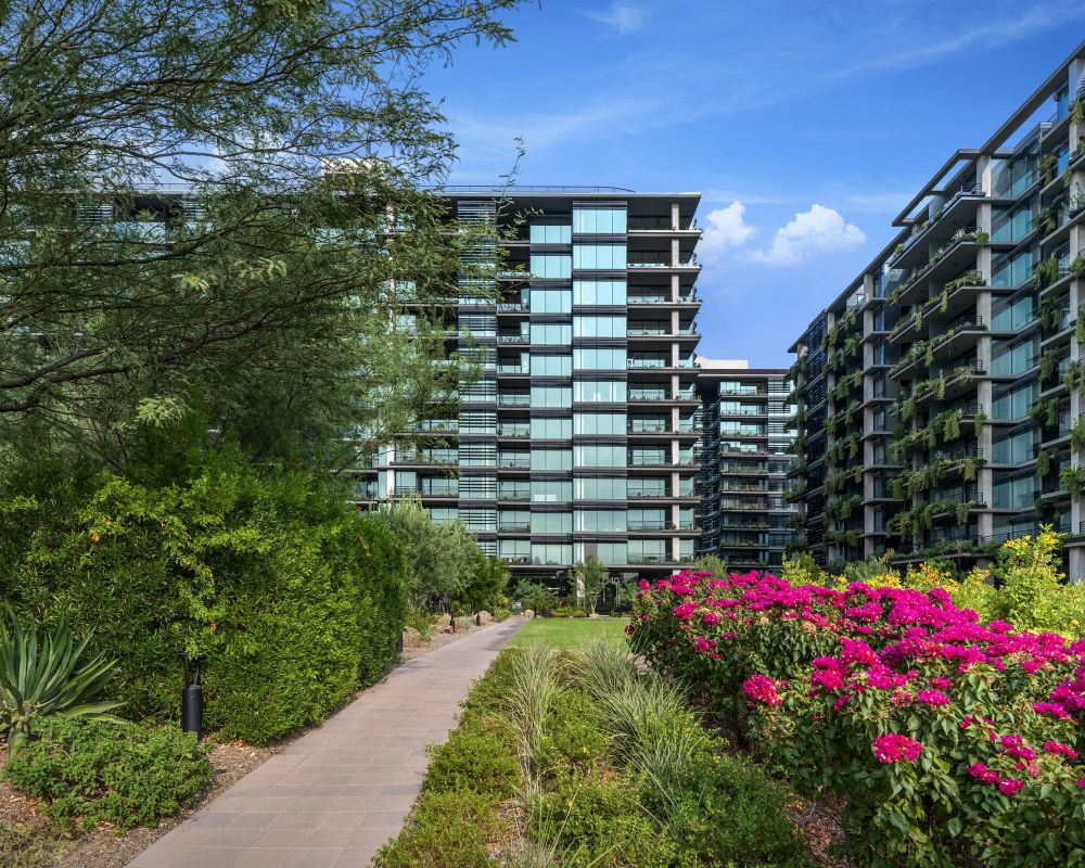 Pathway through lush gardens leading to modern glass apartment buildings under a clear blue sky.