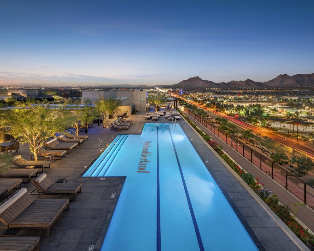 Rooftop infinity pool with lounge chairs overlooking a cityscape and mountains at sunset.