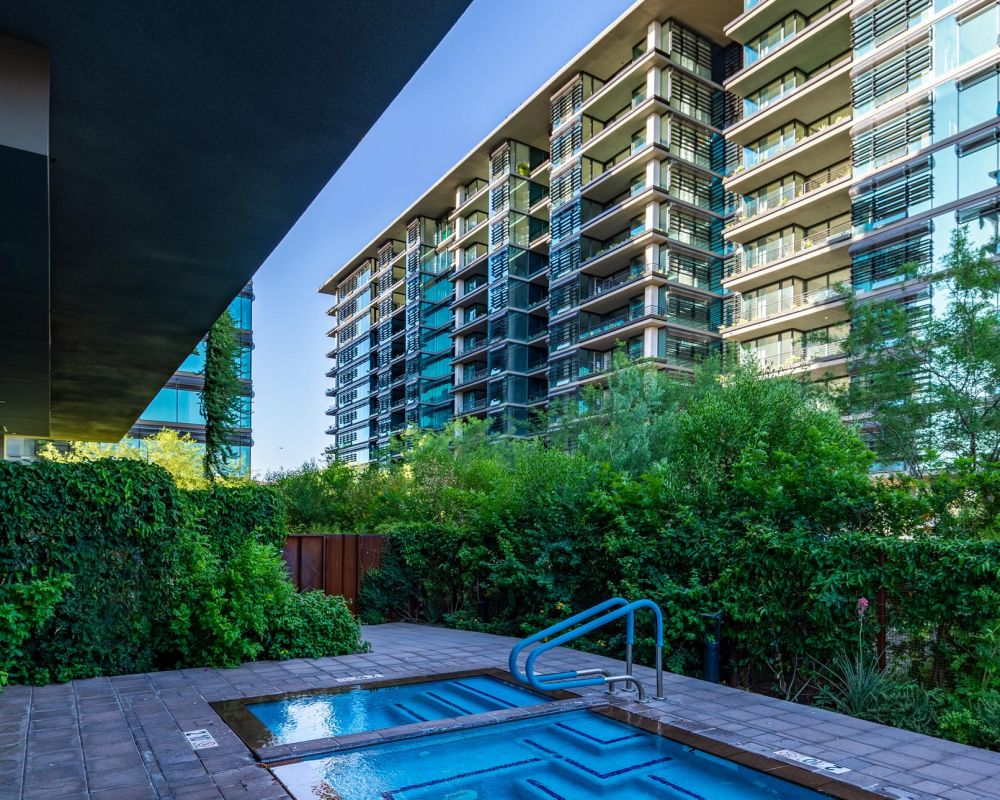 A small outdoor hot tub and pool surrounded by greenery near modern apartment buildings.
