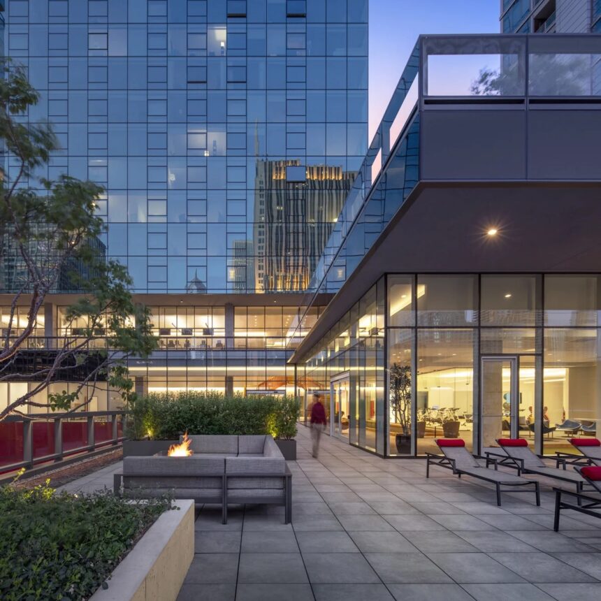Modern outdoor terrace with lounge seating, fire pit, and glass-walled building at dusk.