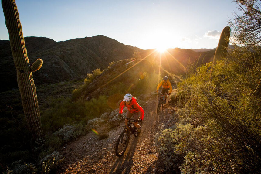 Two people enjoy low-carbon travel, mountain biking a desert trail at sunset among cacti and rugged hills.