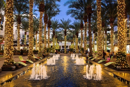 Palm trees wrapped in string lights line a fountain at night in a lively outdoor shopping area.