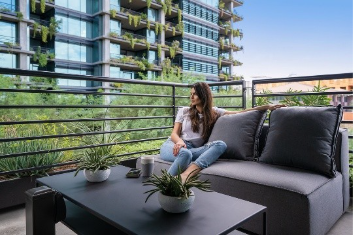 Woman sitting on a balcony sofa, surrounded by plants, with a modern building visible in the background.
