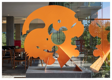 Bright orange abstract sculpture with circular cutouts, showcased indoors near glass windows and wooden chairs, enriching the community art scene.