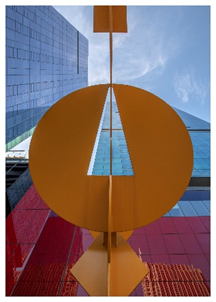 View looking up at a yellow abstract sculpture with tall colorful buildings and blue sky in the background.