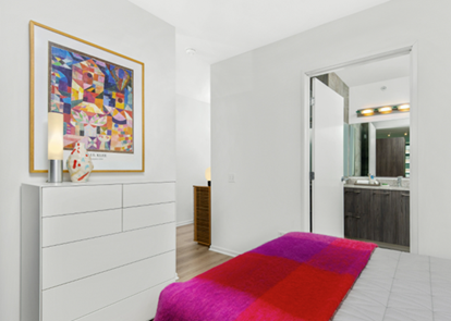 A modern bedroom with a colorful bedspread, white dresser, framed art, and view into a bathroom.