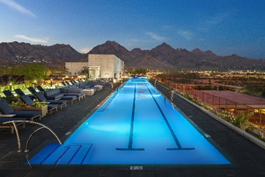Rooftop pool with glowing blue water, lounge chairs, mountain and city views at sunset—luxuriate in signature Optima Sky Decks rooftop amenities.