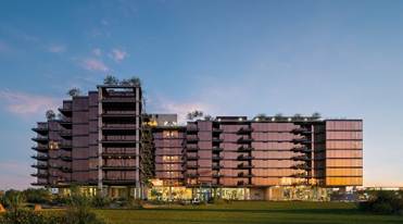 Modern multi-story building with glass exterior, balconies, and rooftop greenery at sunset, viewed from a lawn.