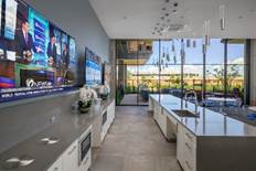 Modern kitchen with large island, two wall-mounted TVs, and floor-to-ceiling windows overlooking a garden.