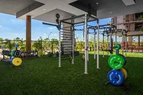Outdoor gym with weight plates, squat racks, and exercise equipment on artificial grass under a covered roof.