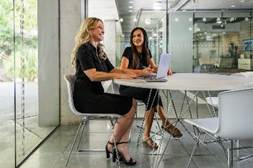 Two women sitting at a round table in a modern office, smiling and working on a laptop together.