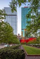 Two modern glass skyscrapers behind green trees and a manicured lawn in an urban park setting.