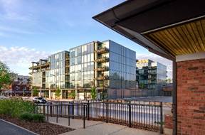 Modern glass apartment building with balconies, next to a brick structure and a fenced sidewalk.