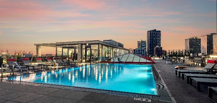 Optima Residents enjoy a rooftop pool at sunset with lounge chairs, city views, and a glass-enclosed structure—choose to rent this urban oasis.